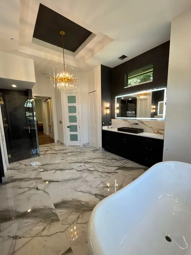 Primary bathroom remodel with a freestanding tub, polished marble-look floor, dark vanity and backlit mirrors under a tray ceiling