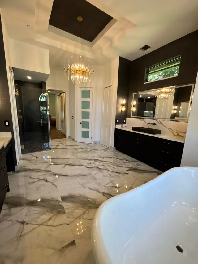 Primary bathroom with a marble floor, a freestanding tub, a black double vanity with backlit mirrors and a crystal chandelier in a tray ceiling