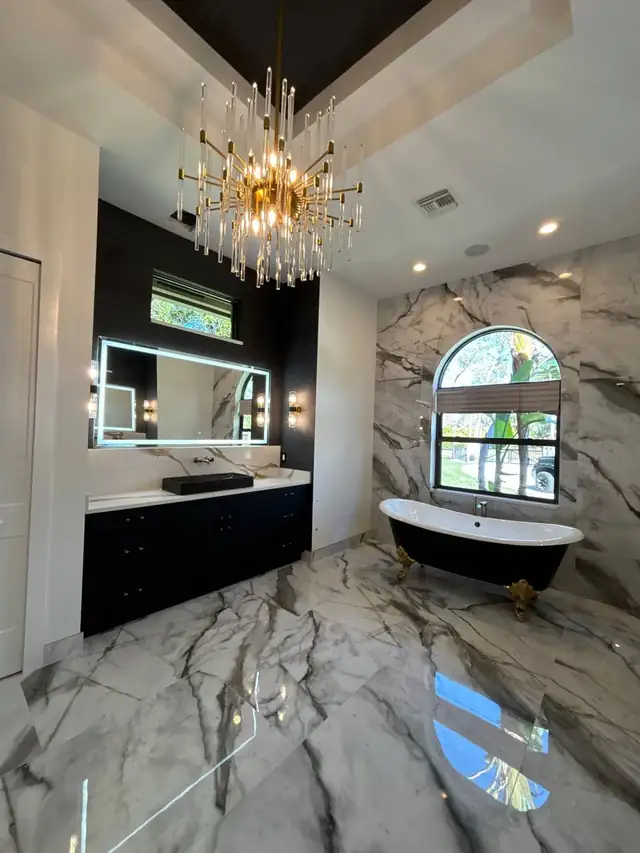 Black freestanding tub under an arched window with a marble feature wall and crystal chandelier